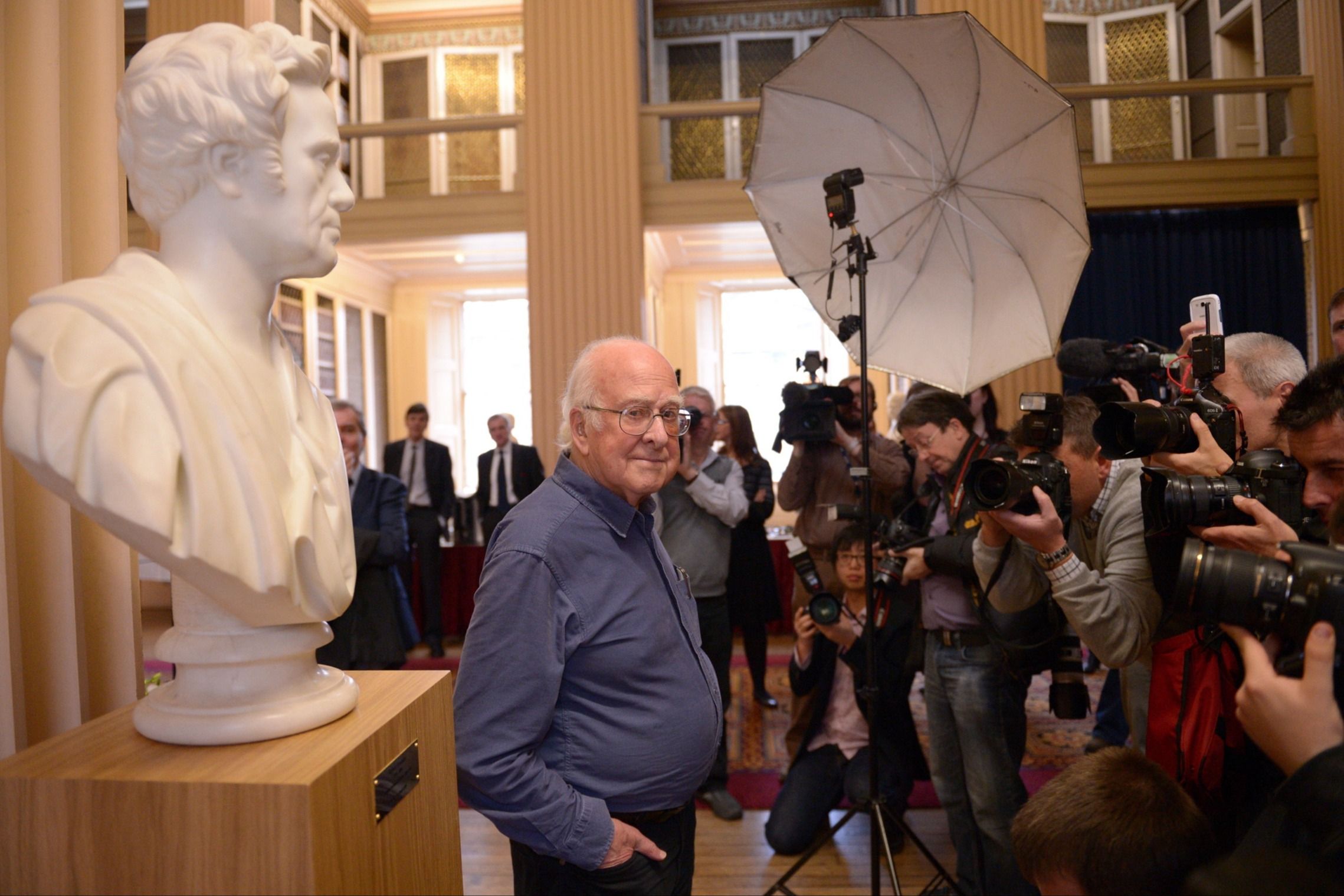 An older man stands in a hall with people holding cameras and TV cameras 