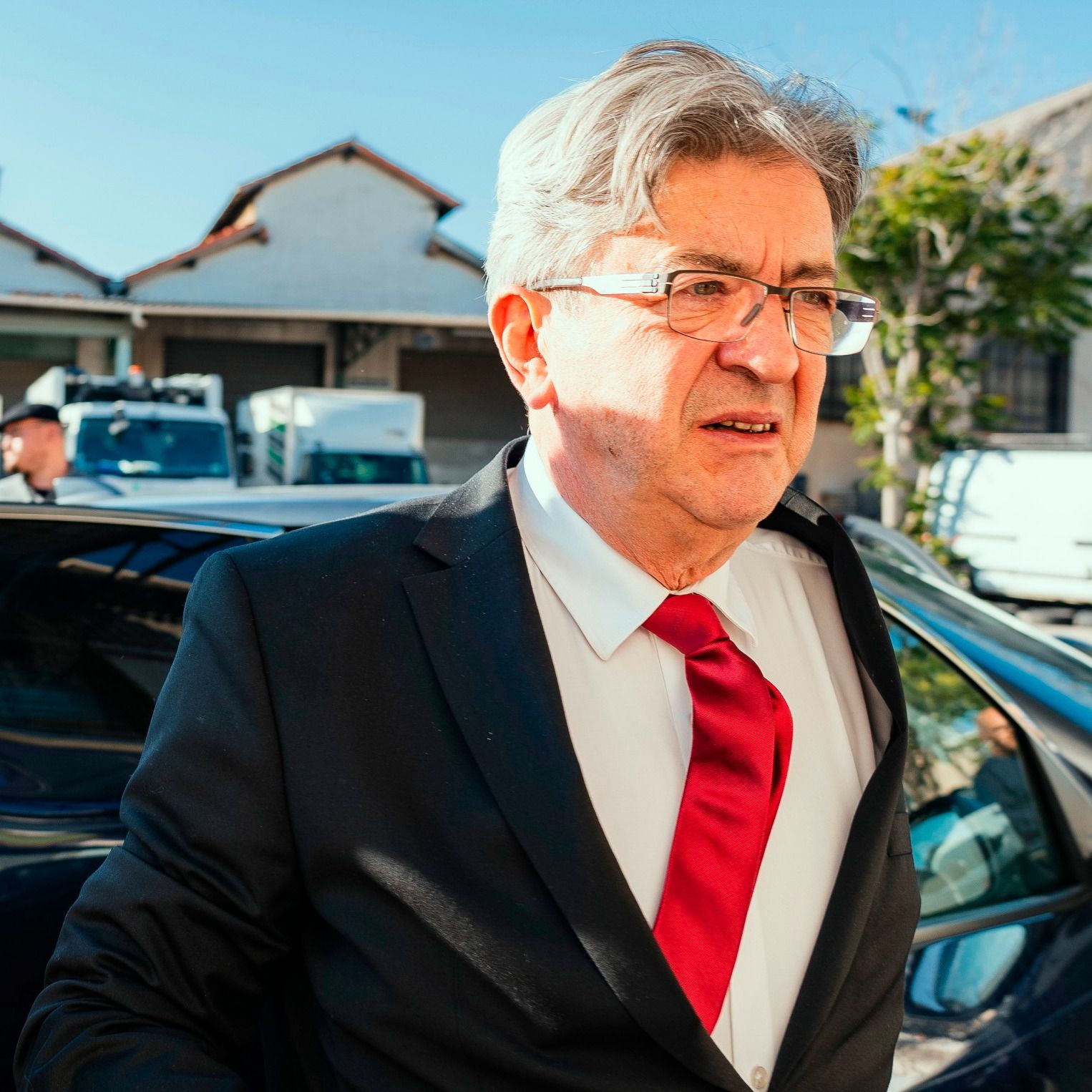 A man in a suit with a red tie stands in front of a car